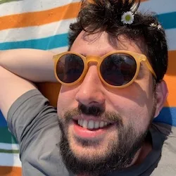 Close up of Kao's face, while he's lying down over a picnic towel, hands behind his head, wearing orange rimmed sunglasses, grey t-shirt and a daisy attached to his dark hair and a big smile surounded by his beard.